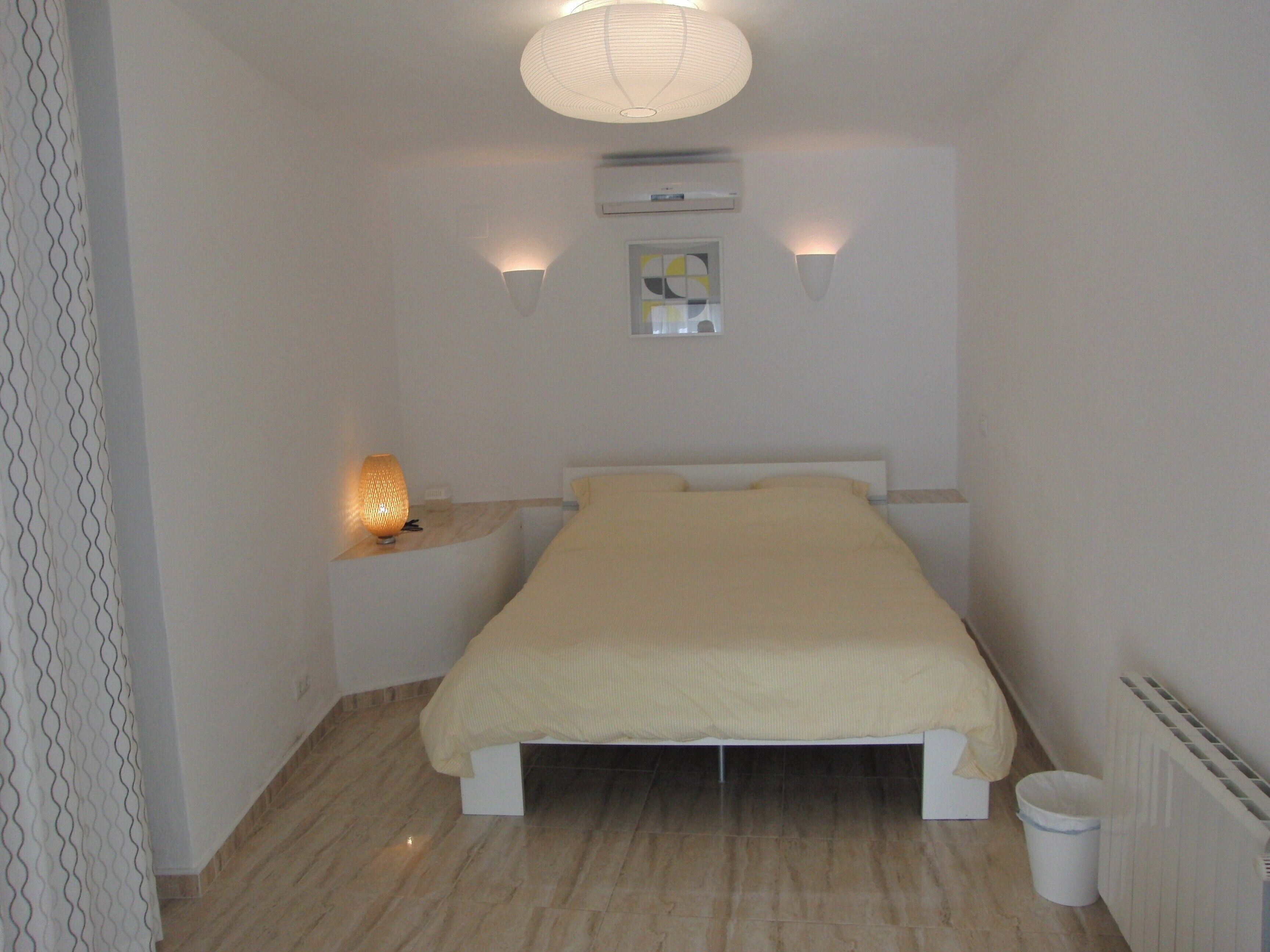 Una habitación pequeña con cama, mesita de noche, lámpara de pared y equipo de aire acondicionado.