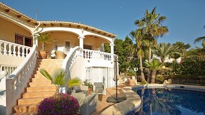 Un espace aménagé au bord de la piscine, avec une piscine aux contours arrondis, un escalier menant à un porche couvert et un palmier en arrière-plan.