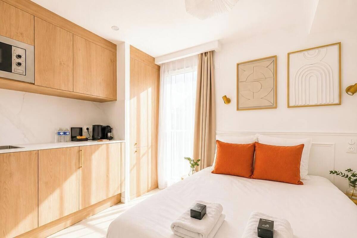 A modern hotel room with a kitchen area, a bed with white linens and orange pillows, and two framed artworks on the wall.