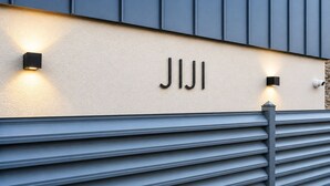 Een gevel met de naam 'JIJI' verlicht in zwarte letters, een geribbelde beige muur en een modern ontwerp met verticale panelen en een schuin dak.