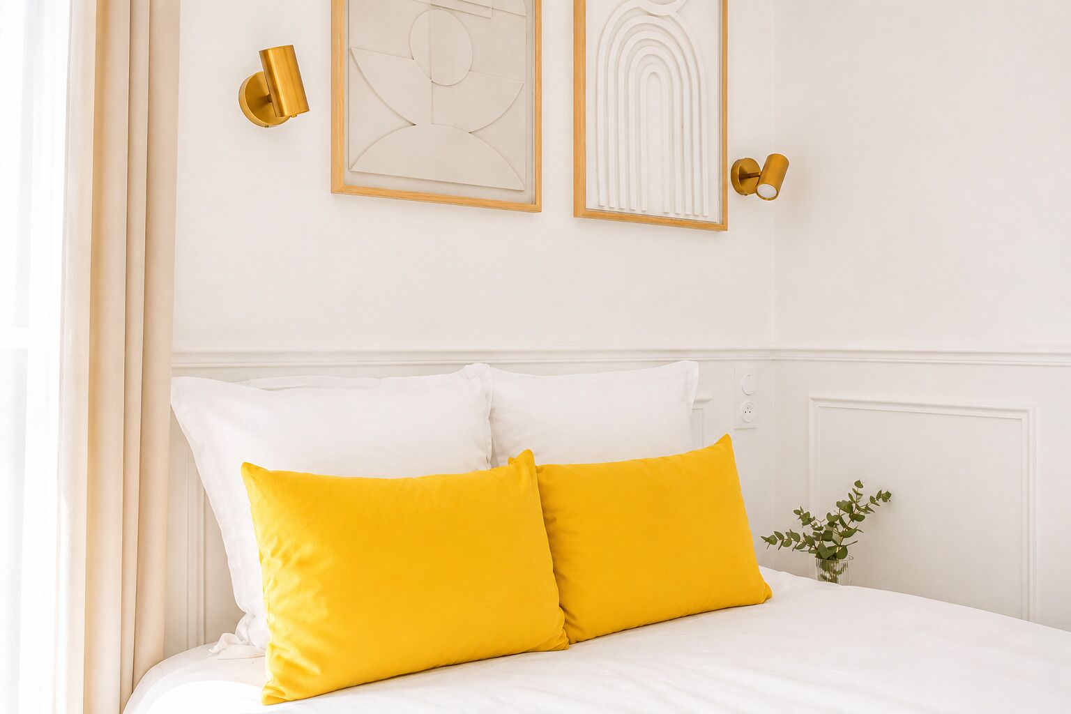 Una cama con almohadas blancas y amarillas, un aplique de pared y una obra de arte enmarcada.