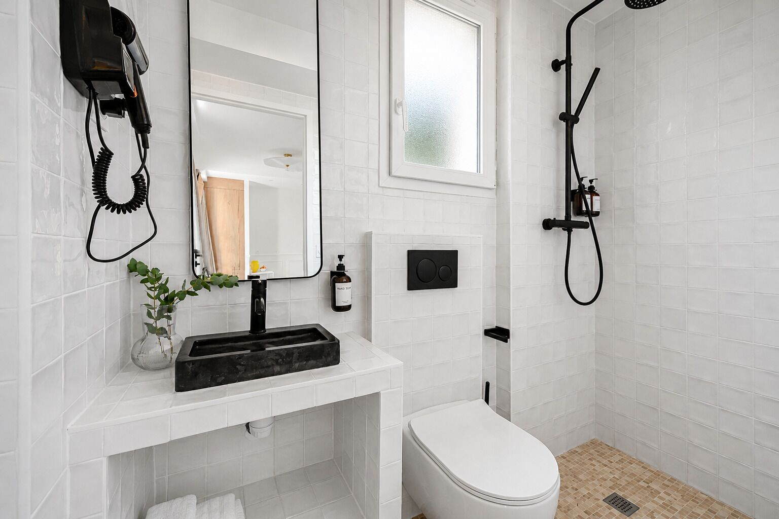 Un bagno moderno con parete rivestita di piastrelle bianche, lavandino nero, un water, una doccia e una finestra.