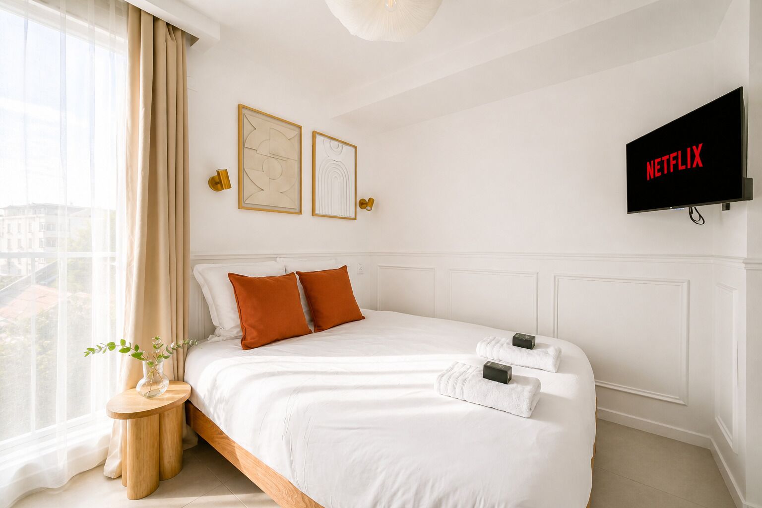 Una habitación de hotel con cama, mesita de noche con un florero, televisor en la pared y ventana con cortinas.