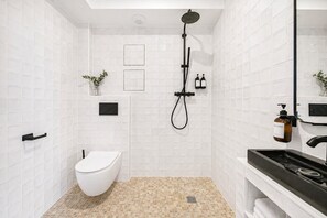 Un baño moderno con paredes de azulejos blancos, inodoro blanco, ducha con cabezal negro, lavabo negro y una pequeña planta en maceta.