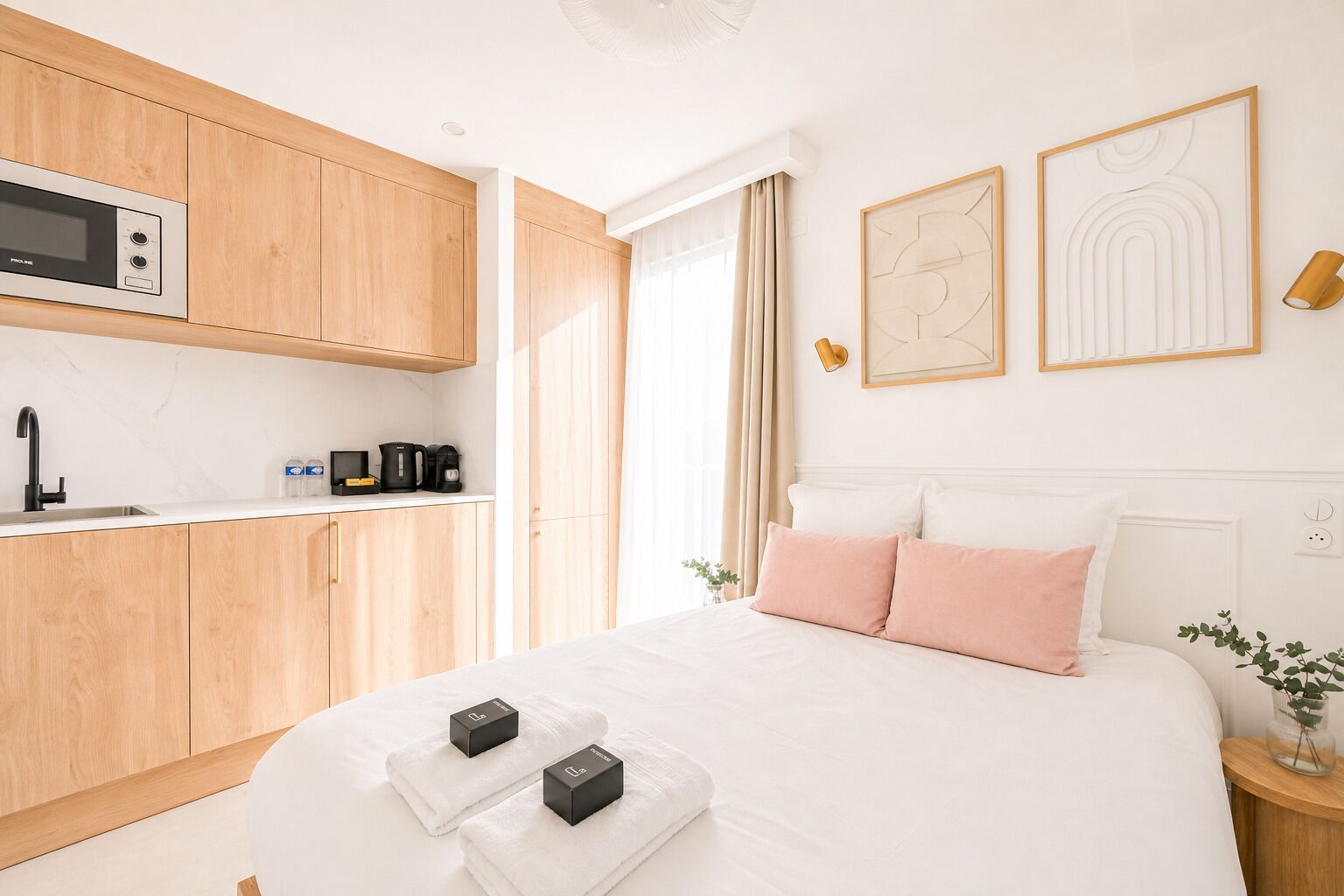 Ein modernes Hotelzimmer mit Küchenzeile, einem Bett mit weißer und rosafarbener Bettwäsche und zwei an der Wand befestigten Kunstwerken.