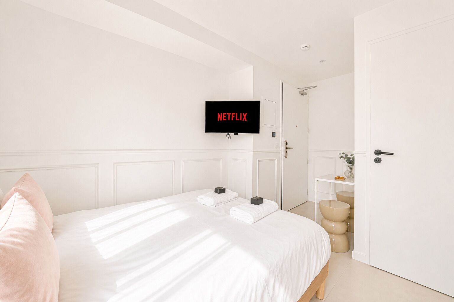 A hotel room with a bed, a TV mounted on the wall, a door, and a small table with a vase.