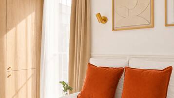 Quarto com cama, cortinas bege, luminária fixada na parede e obra de arte abstrata emoldurada.
