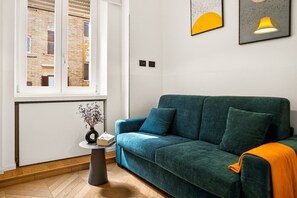 Una sala moderna con un sofá verde azulado, una mesita auxiliar con un florero y una ventana con vista a un edificio de ladrillo.