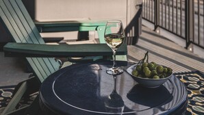 A table with a bowl of green olives and a glass of white wine, a green chair, and a white cooler on a balcony with a metal railing and a view of hills.