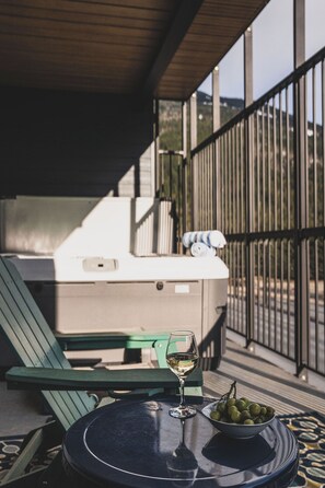 A table with a bowl of green olives and a glass of white wine, a green chair, and a white cooler on a balcony with a metal railing and a view of hills.