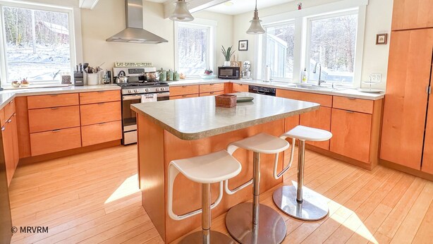 Une cuisine moderne avec des meubles en bois, un îlot central doté de tabourets, des appareils en acier inoxydable et de grandes fenêtres offrant une vue sur l’extérieur.