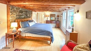 Une chambre confortable avec un mur en pierre, des poutres apparentes en bois au plafond, un lit avec une literie bleue, des lampes de chevet, une commode et une fenêtre offrant une belle vue.