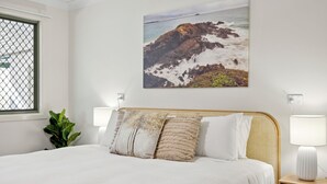 A neatly made bed with a white duvet, two bedside lamps, and a framed coastal landscape painting above it.