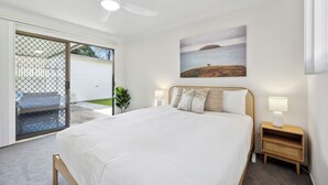 A bedroom with a large bed, a ceiling fan, a nightstand with a lamp, and a view of the sea through a window.
