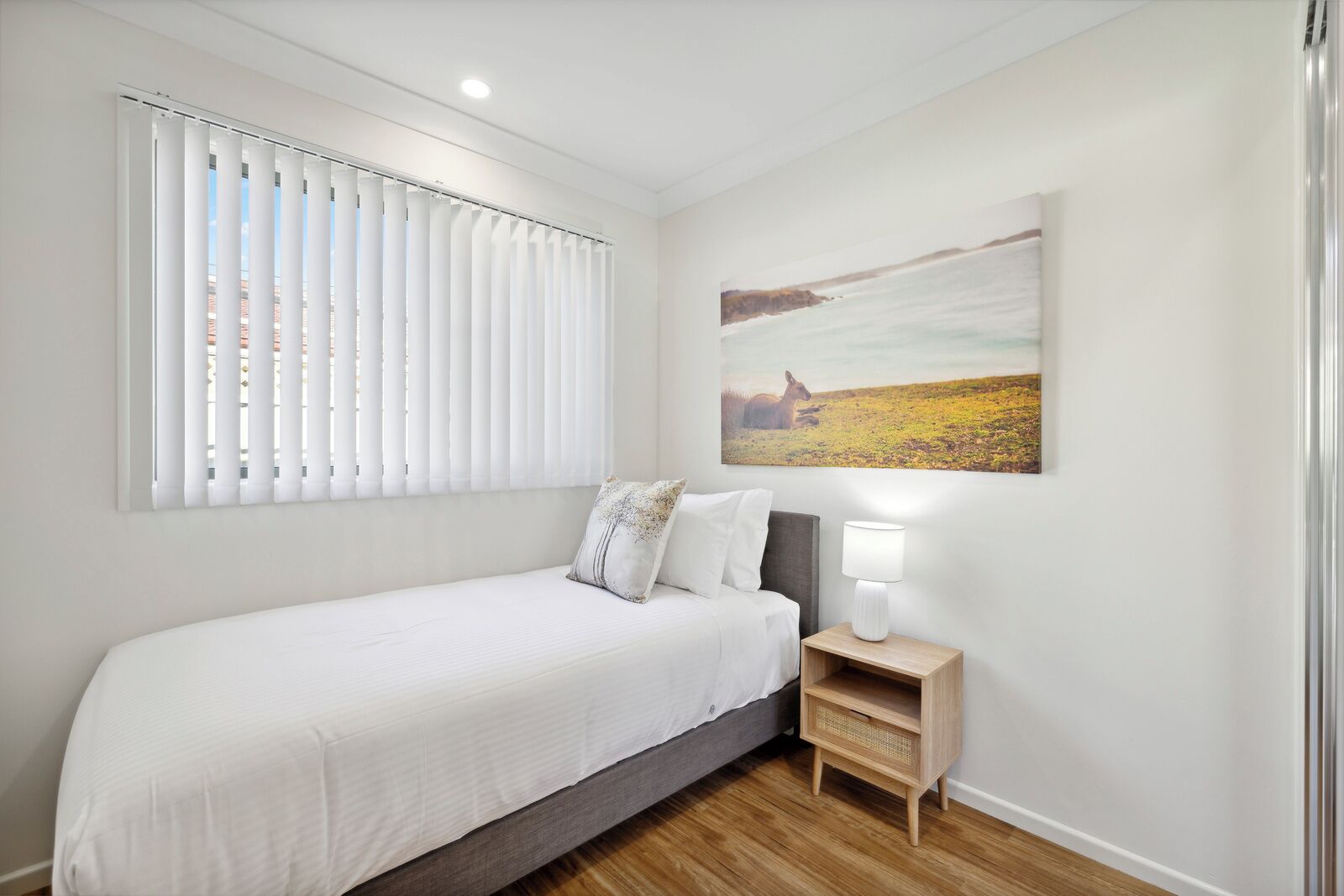 Een slaapkamer met een bed, nachtkastje, lamp, raam met jaloezieën en een schilderij aan de muur.