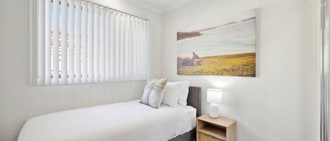 Een slaapkamer met een bed, nachtkastje, lamp, raam met jaloezieën en een schilderij aan de muur.