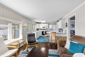 Enjoy a cozy living space with a view of the water through large windows, featuring a seating area and a kitchenette.