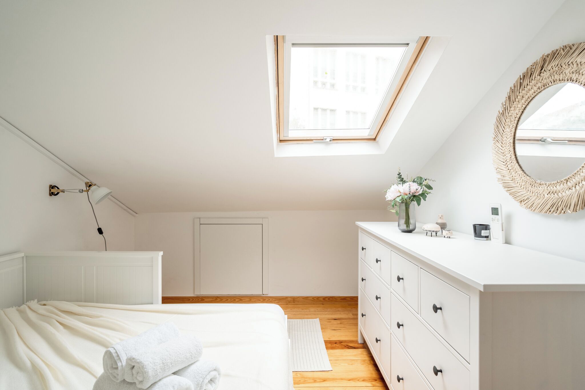 Een slaapkamer met dakraam, een bed met witte lakens, een kapstok en een ronde spiegel.