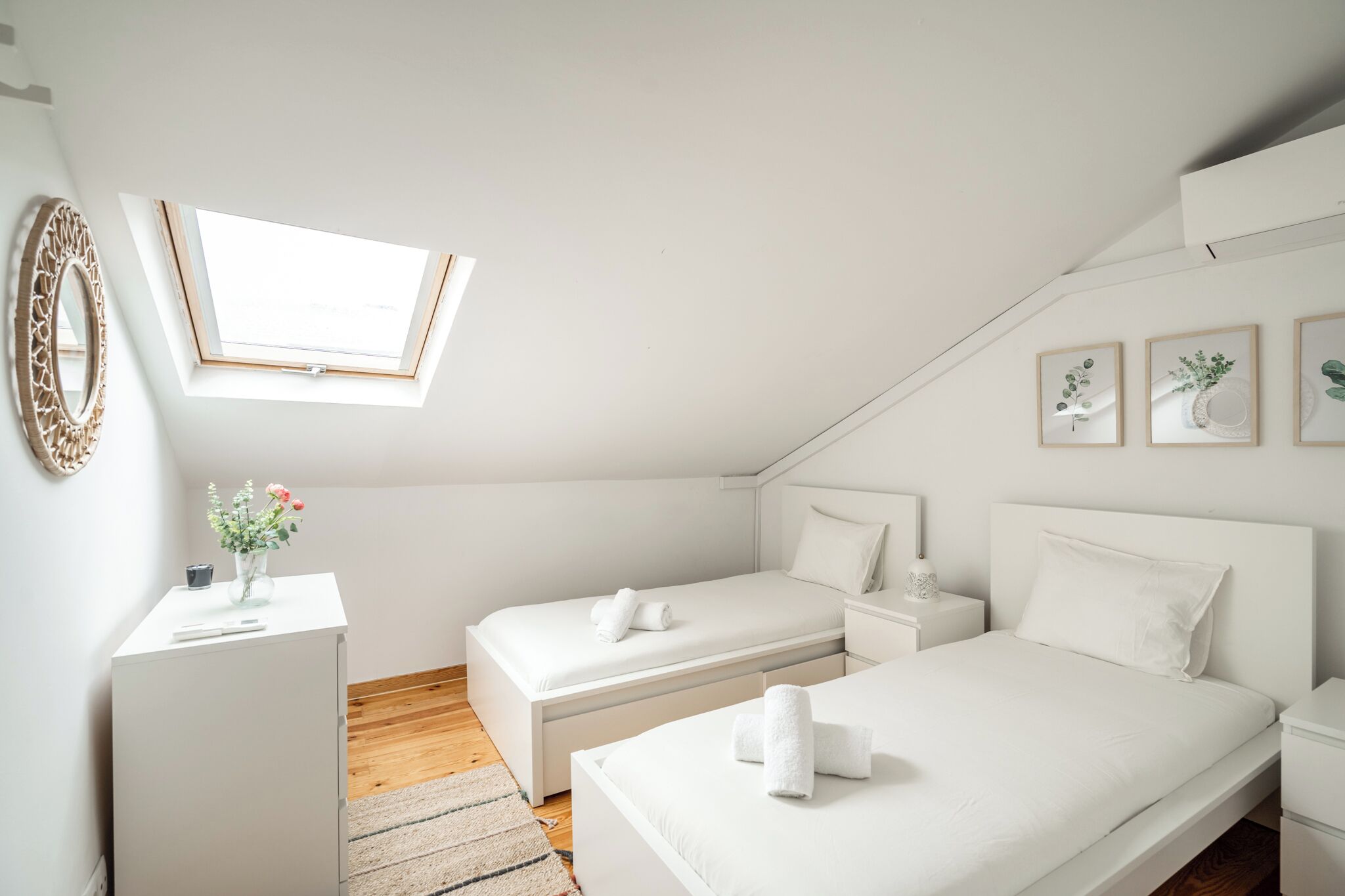 A modern, white-themed bedroom with two beds, a skylight, and framed botanical prints on the wall.