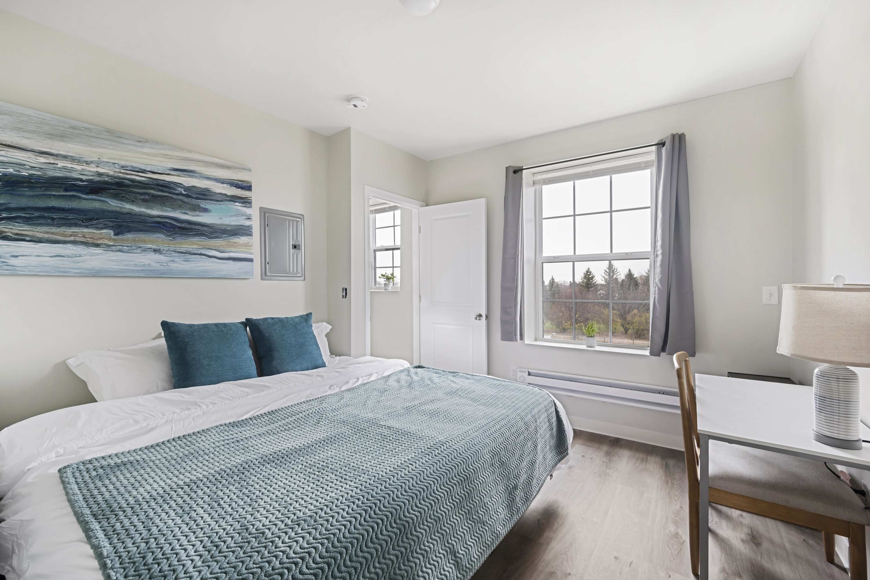 Une chambre à coucher avec un lit, un bureau, une chaise, une fenêtre avec des rideaux et un tableau au mur.