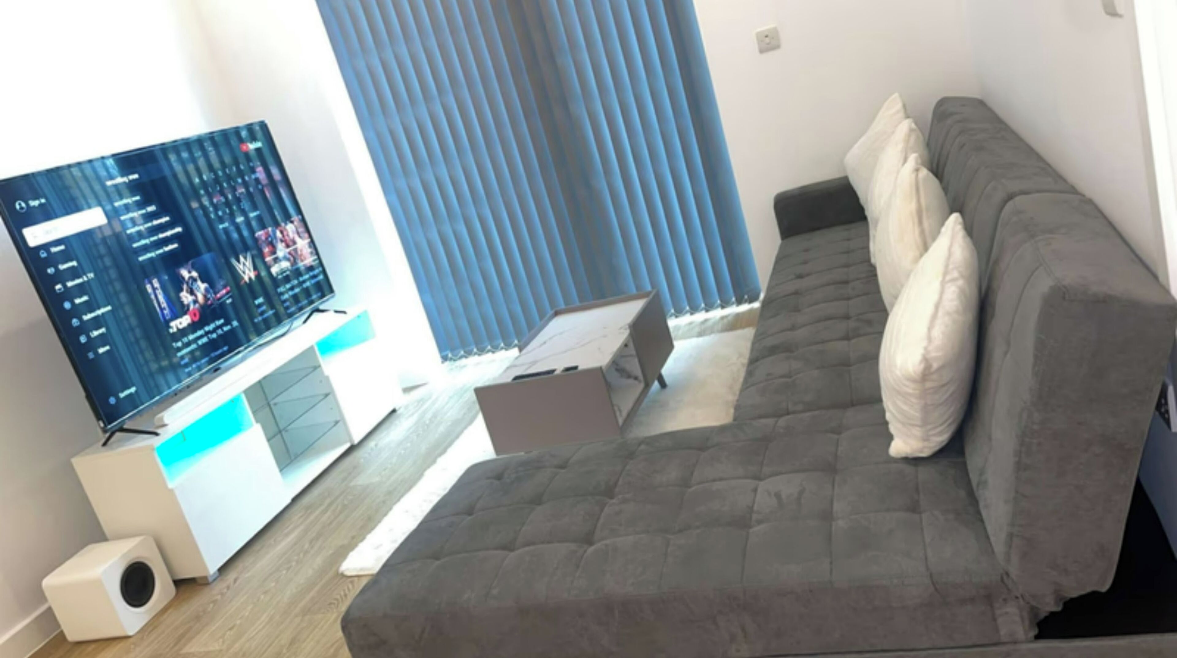 A modern bedroom with a grey sofa, a flat-screen TV displaying a wrestling show, and a window with blue blinds.