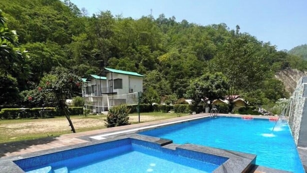 A swimming pool with a clear blue water, surrounded by lush greenery and a building with a green roof. - Aspen Adventures Camp by Aspen (Rishikesh)
