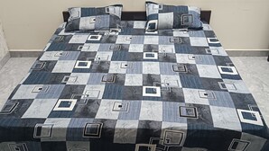 A bed with a checkered bedspread and two matching pillows in a room with a tiled floor and a plain wall. - Hariom Home Stay By GRB (Faizabad)