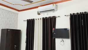 A room with a ceiling fan, an air conditioning unit, a black cabinet, and two sets of curtains. - Hariom Home Stay By GRB (Faizabad)