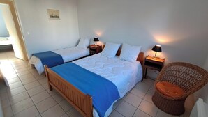 Una habitación de hotel con dos camas individuales, cabecera de madera, una silla de mimbre y una mesita con lámpara.