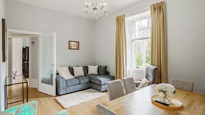 A modern living room with a sectional sofa, a dining table with chairs, a chandelier, and a view of a cityscape through a window with curtains. - Spacious City Apartment - Dunfermline (Dunfermline)