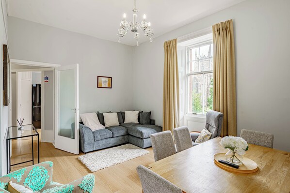 A modern living room with a sectional sofa, a dining table with chairs, a chandelier, and a view of a cityscape through a window with curtains.
