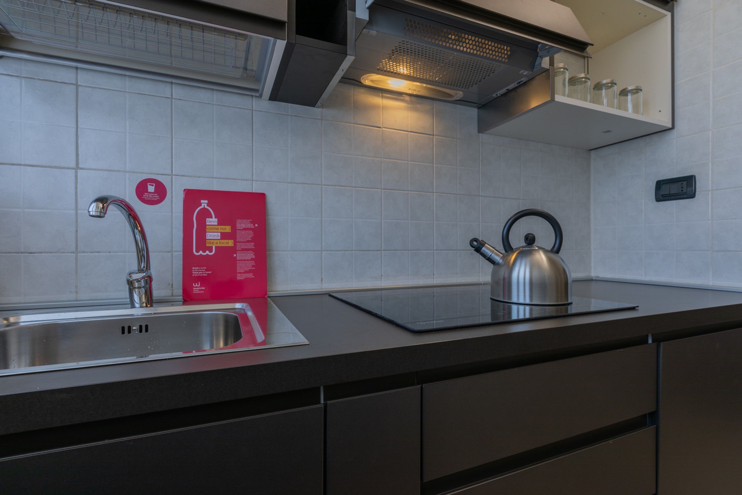 A modern kitchen with a stainless steel sink, a kettle, and a red informational card on the counter.