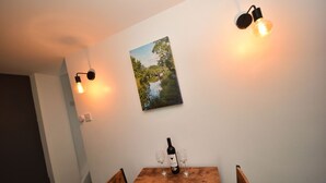 Zona pranzo con tavolo in legno, due bicchieri da vino e una bottiglia. Due lampade a parete e un quadro appeso al muro.