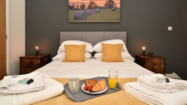 Un letto rifatto con cuscini, un vassoio con croissant, fragole e succo d'arancia, e un quadro con balle di fieno sulla parete.