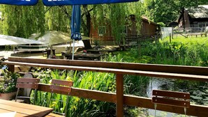 A wooden deck with benches overlooking a pond, surrounded by lush greenery and a rustic building in the background. - Sterne Luxus Heidesonne (Jesteburg)