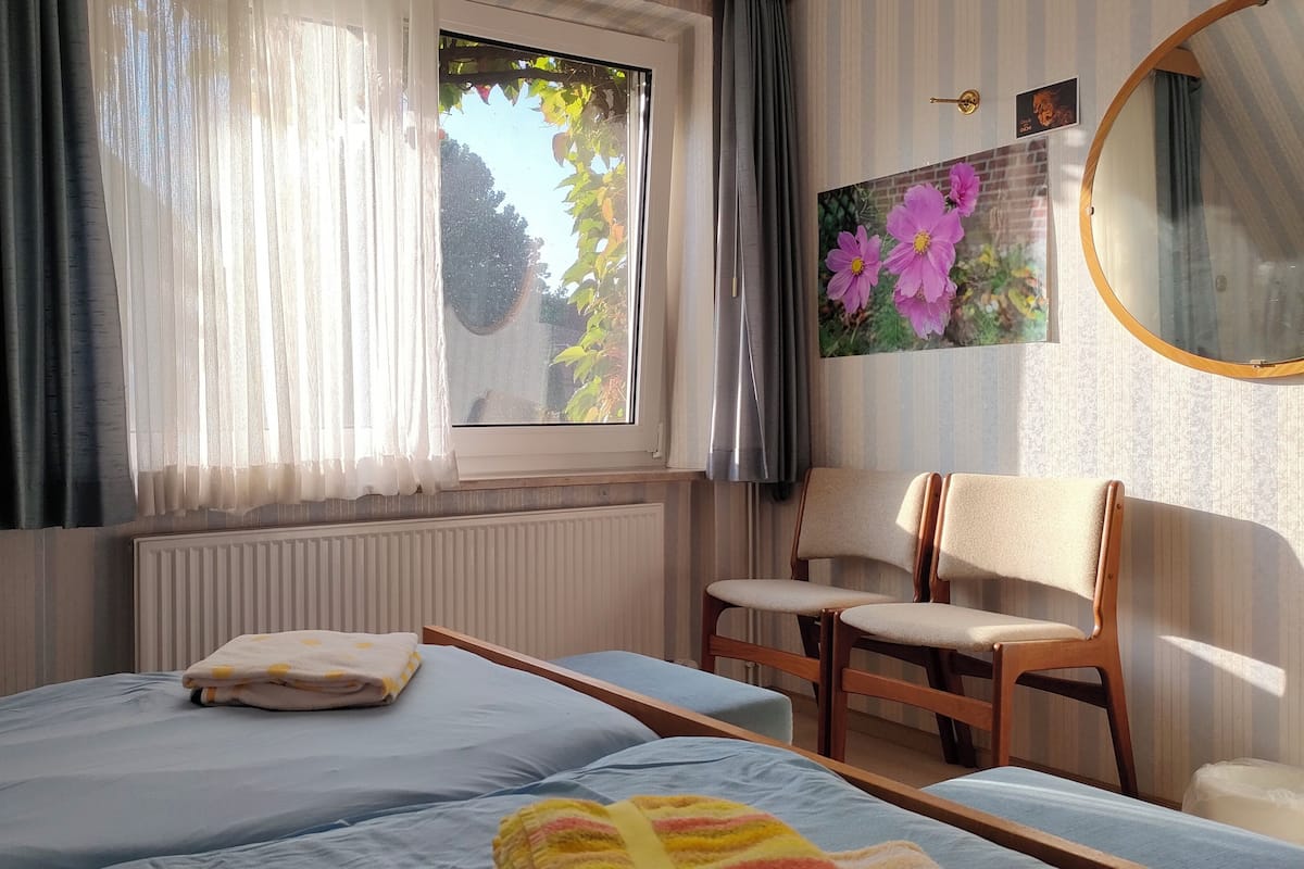 A bedroom with a bed, a chair, a window with curtains, a radiator, and a wall-mounted mirror.