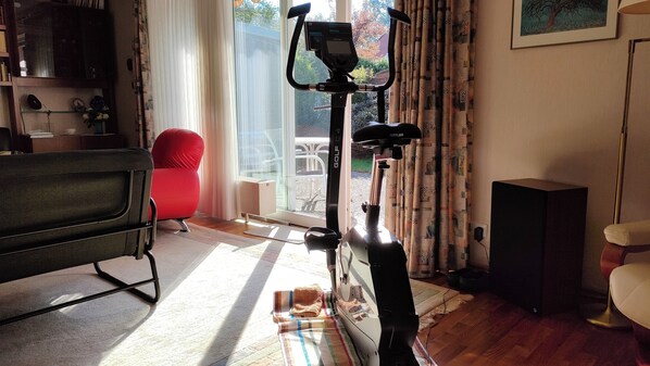 Una habitación con una bicicleta estática, una silla roja, un altavoz negro y una ventana con cortinas.