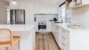 A modern kitchen with white cabinetry, a stainless steel refrigerator, an oven, and a dining area with a wooden chair.