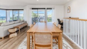 A spacious room with a large wooden dining table, white walls, and a balcony with a view of the sea.