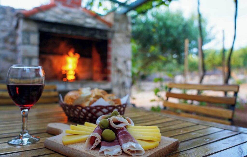 A wooden table with a glass of red wine, a wooden cutting board with cheese, olives, and prosciutto, and a basket of bread.