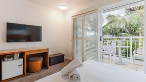 Habitación de hotel con cama, televisor, escritorio pequeño y balcón con vista a palmeras.