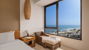Un dormitorio con un ventanal grande, una cama, una mesita de noche, una silla de mimbre y vista al océano.
