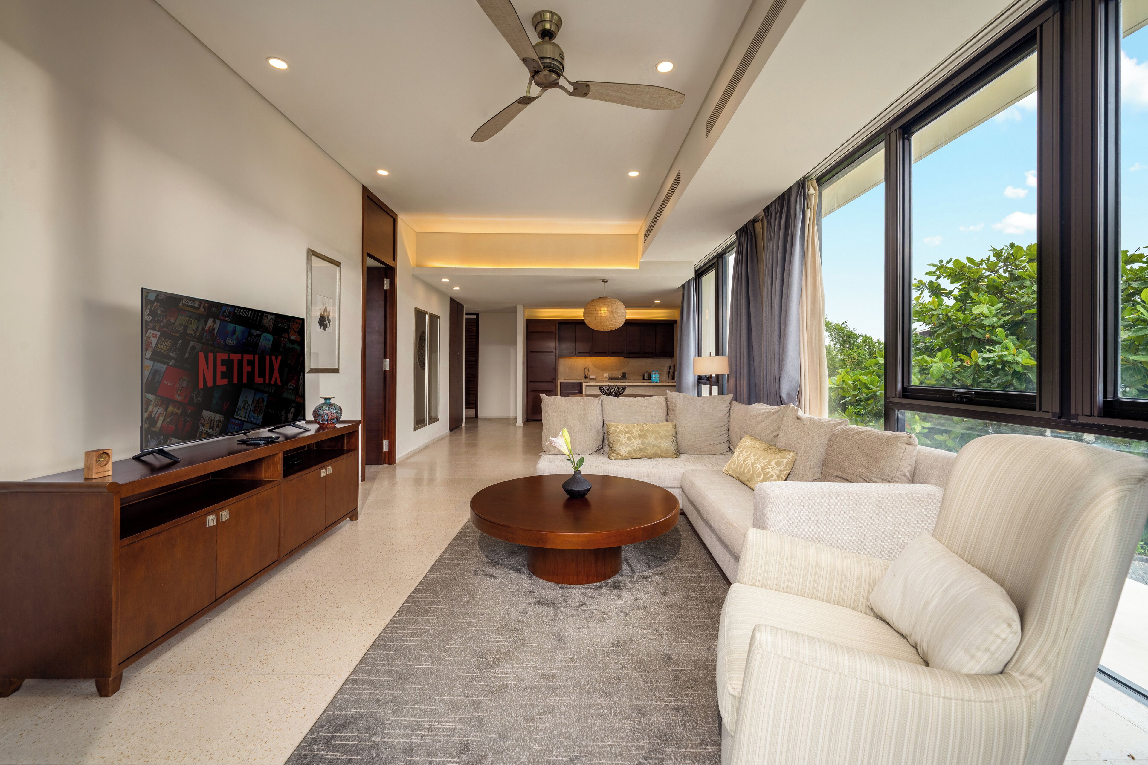 A modern living room with a large flat-screen TV, a wooden entertainment unit, a round coffee table, a beige sofa, and a view of the outdoors through large windows.