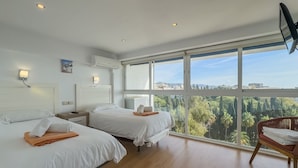 A hotel room with two beds, a large window with a city view, and a flat-screen TV mounted on the wall.