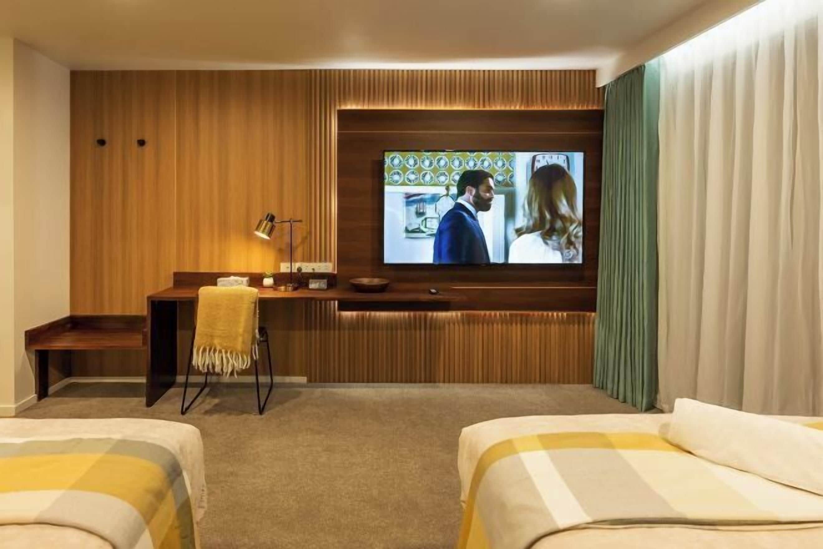 A hotel room with a wooden panel wall, a television mounted above a desk, a bed with a yellow and white striped bedspread, and a window with curtains.