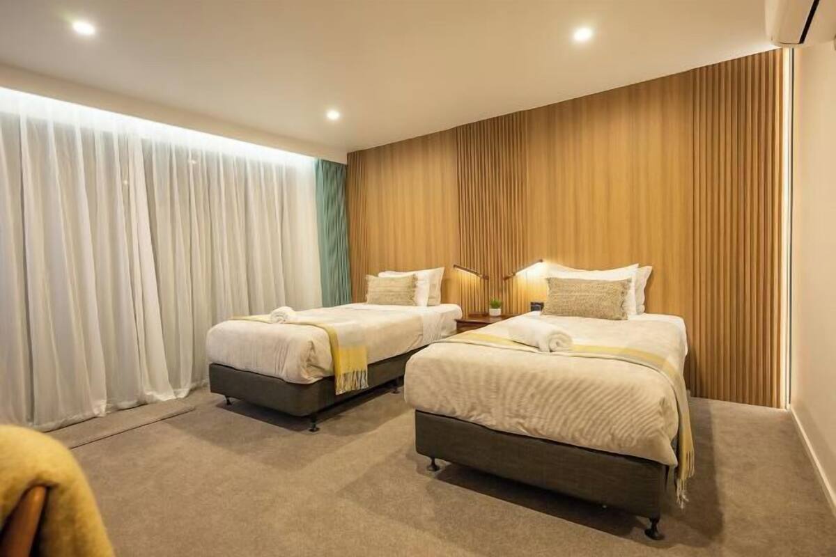 Une chambre d’hôtel avec deux lits, un mur décoratif en bois et une grande fenêtre avec des rideaux légers.