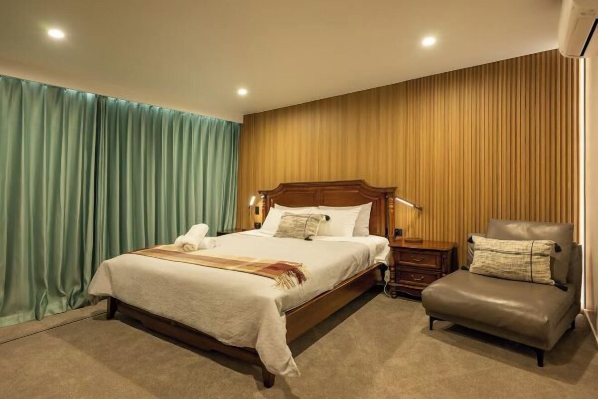A hotel room with a large bed, a wooden headboard, a nightstand, a chair, and a wooden wall panel.