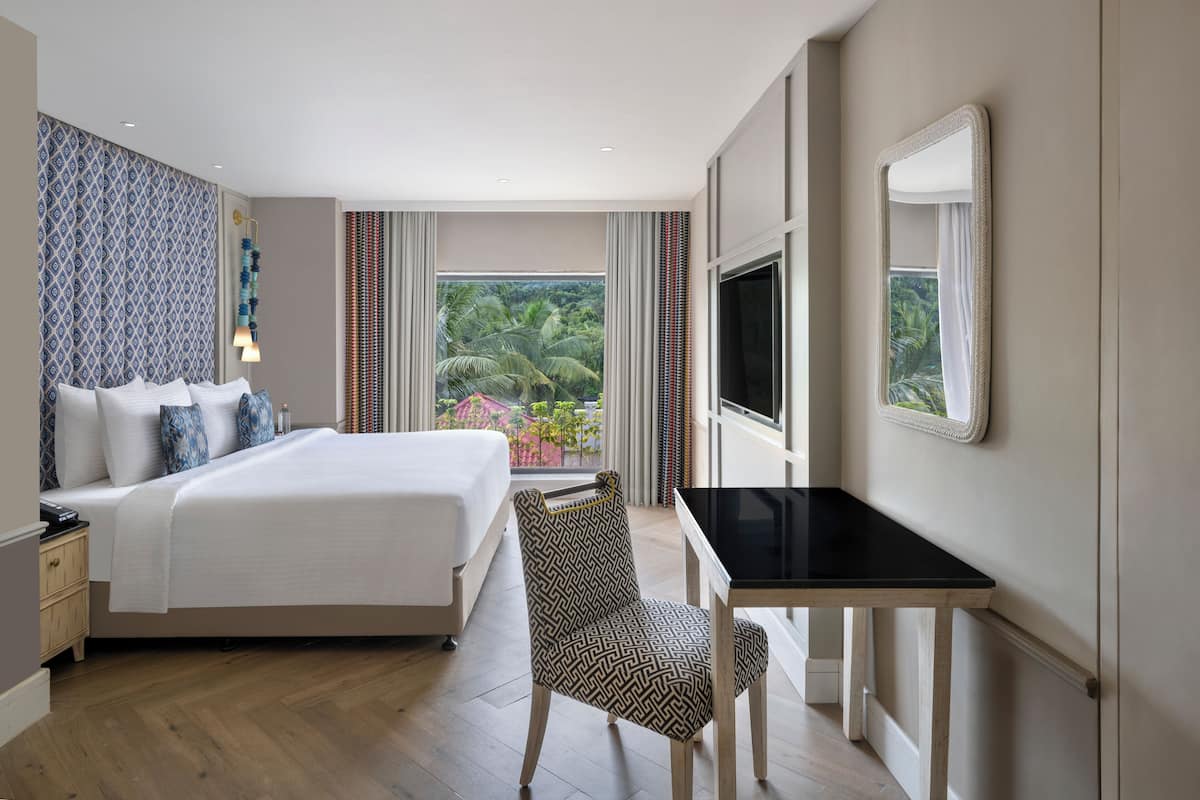 A hotel room with a bed, a desk, a chair, a TV, and a large window with a view of palm trees.