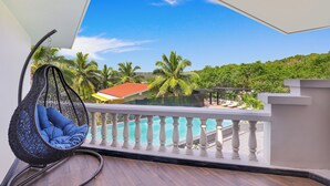 Un balcón con una silla colgante, una piscina y vistas a palmeras y una colina.