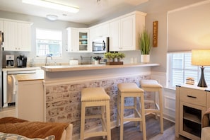 Une cuisine avec des meubles blancs, un mur décoratif en briques, une machine à café et deux tabourets.